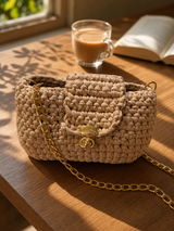 Sona Flap Bag in biscuit tan crochet with gold chain, on a wooden table beside a cup of chai